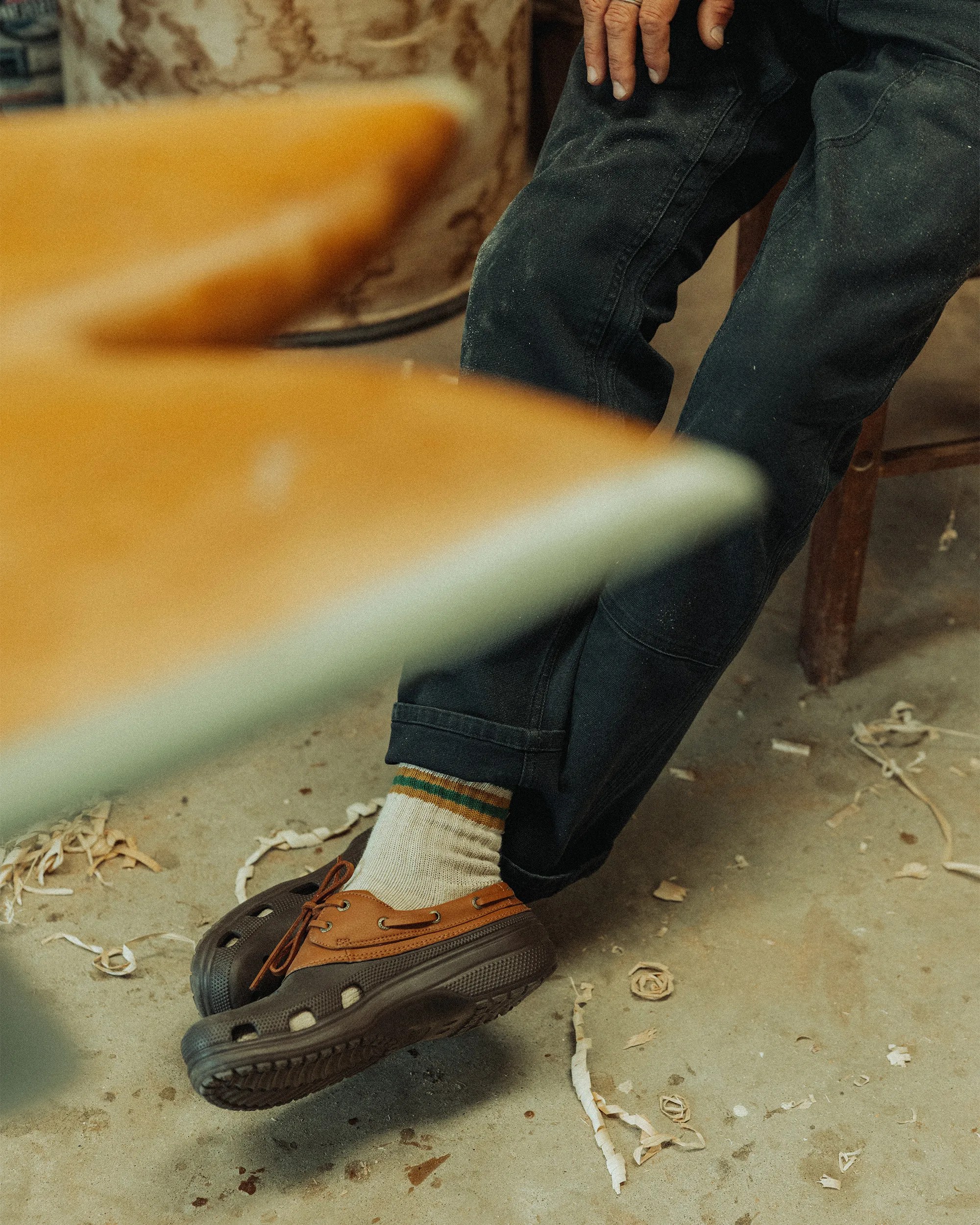 A person wearing black pants, white socks with green and yellow stripes, and brown slip-on shoes with black soles is sitting on a wooden chair. The floor is concrete with wood shavings scattered around. The person's hand rests on their knee. In the foreground, there is a blurred yellow object, possibly a surfboard.