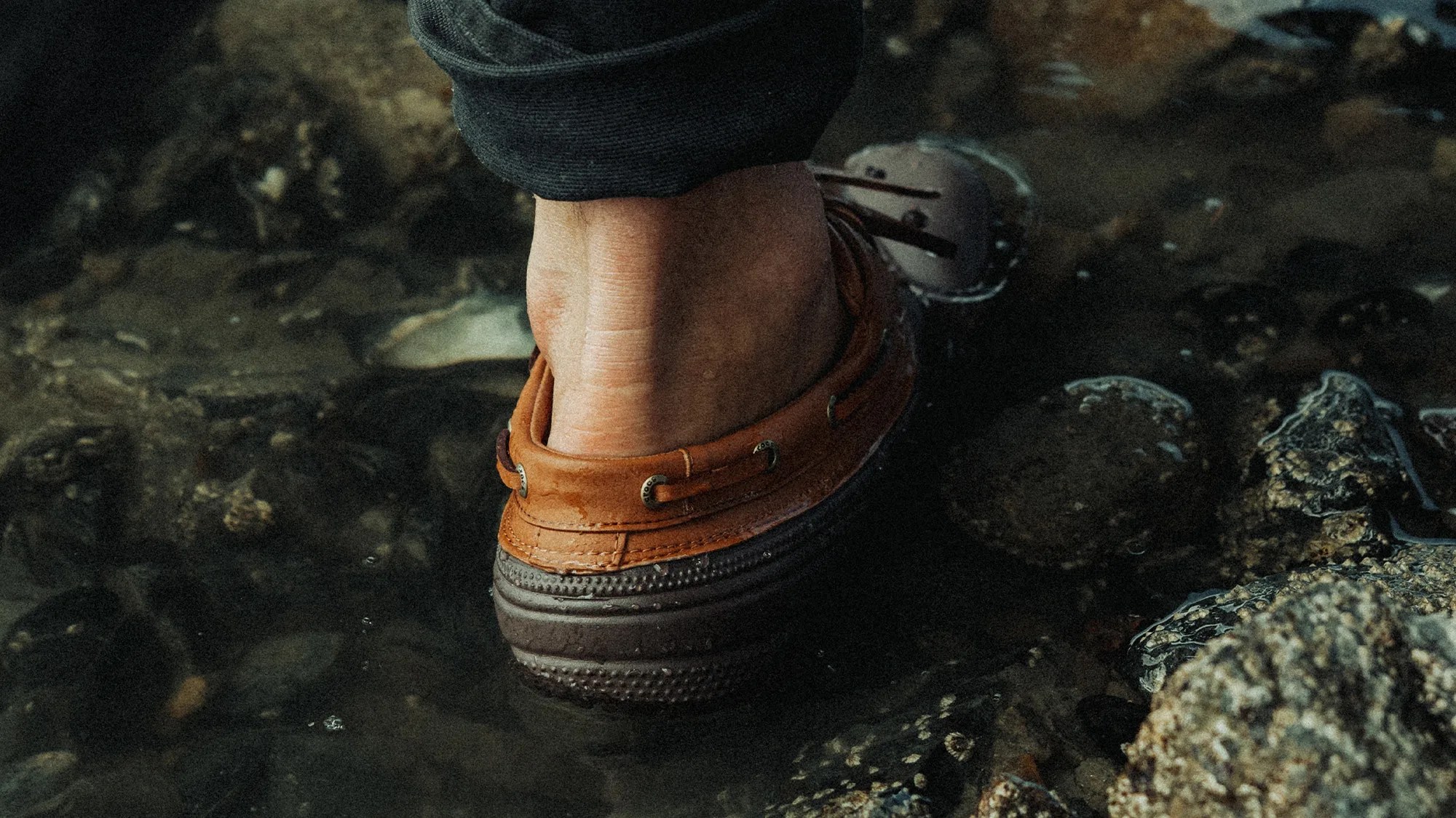 A close-up of a person's foot wearing a brown leather boat shoe with dark soles, partially submerged in shallow water with visible rocks and pebbles underneath. The person is wearing dark pants rolled up slightly above the ankle.