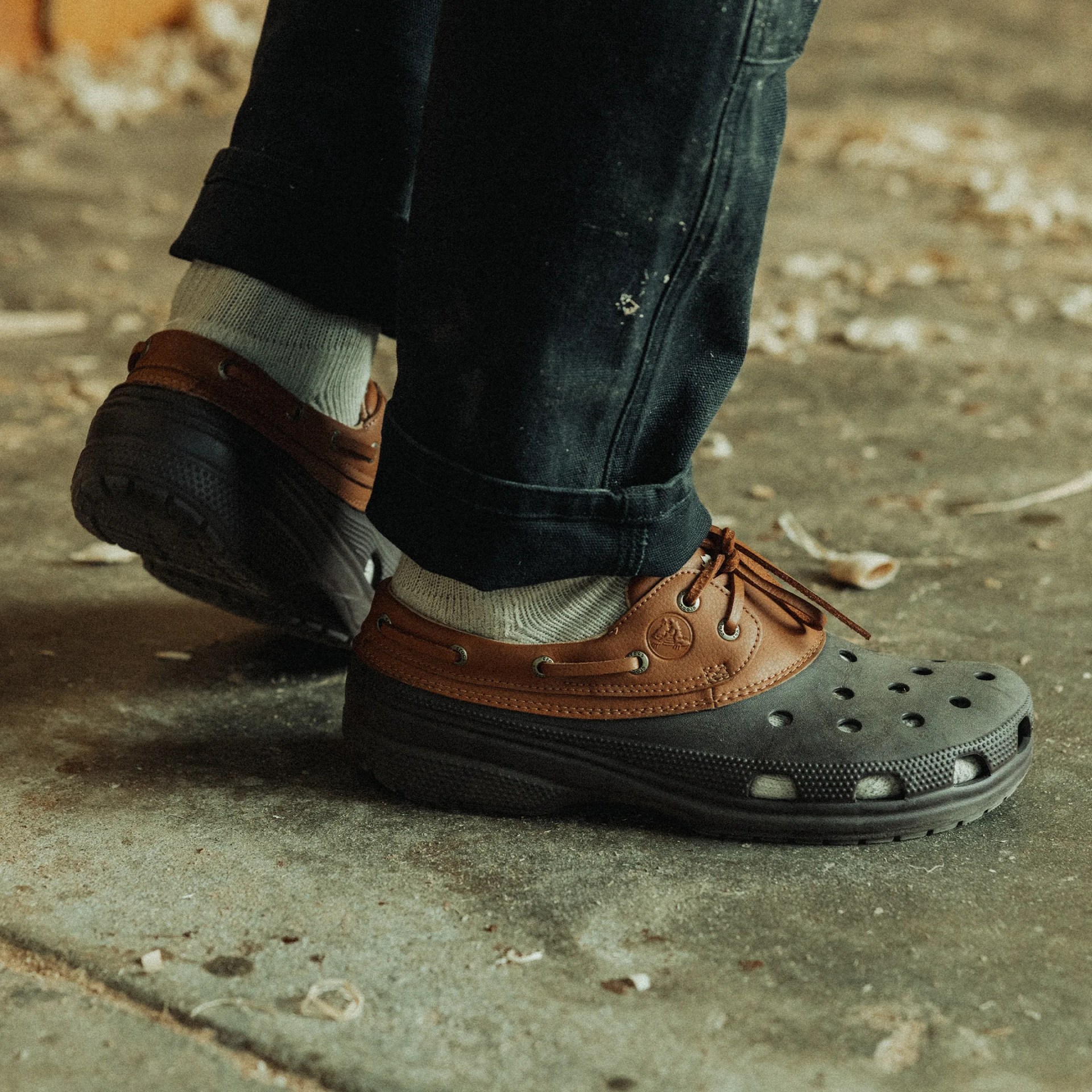 Brown and black slip-on shoes with holes on the toe area, worn with light-colored socks and black pants, standing on a rough concrete floor.