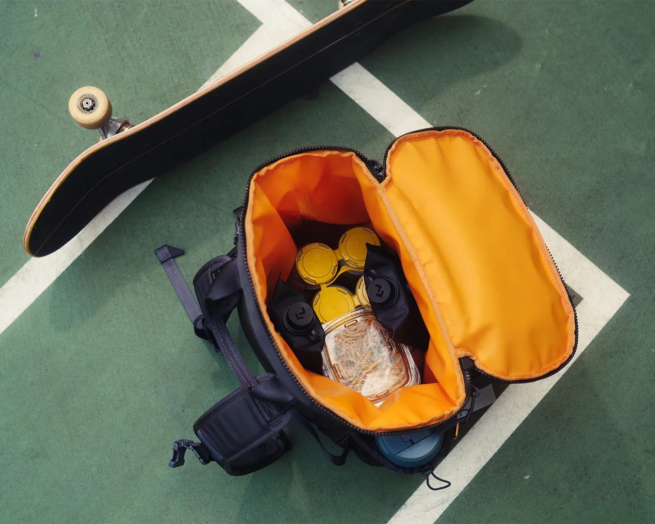 Open black and orange insulated cooler bag containing several yellow-lidded containers and a clear plastic container with food, placed on a green sports court next to a skateboard.