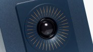 Close-up of a black speaker driver with a central dome surrounded by evenly spaced gold radial lines on a dark blue textured surface.