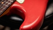 Close-up of a red electric guitar body showing the smooth, glossy finish and part of the neck with metal frets and strings. The guitar rests on a black surface.