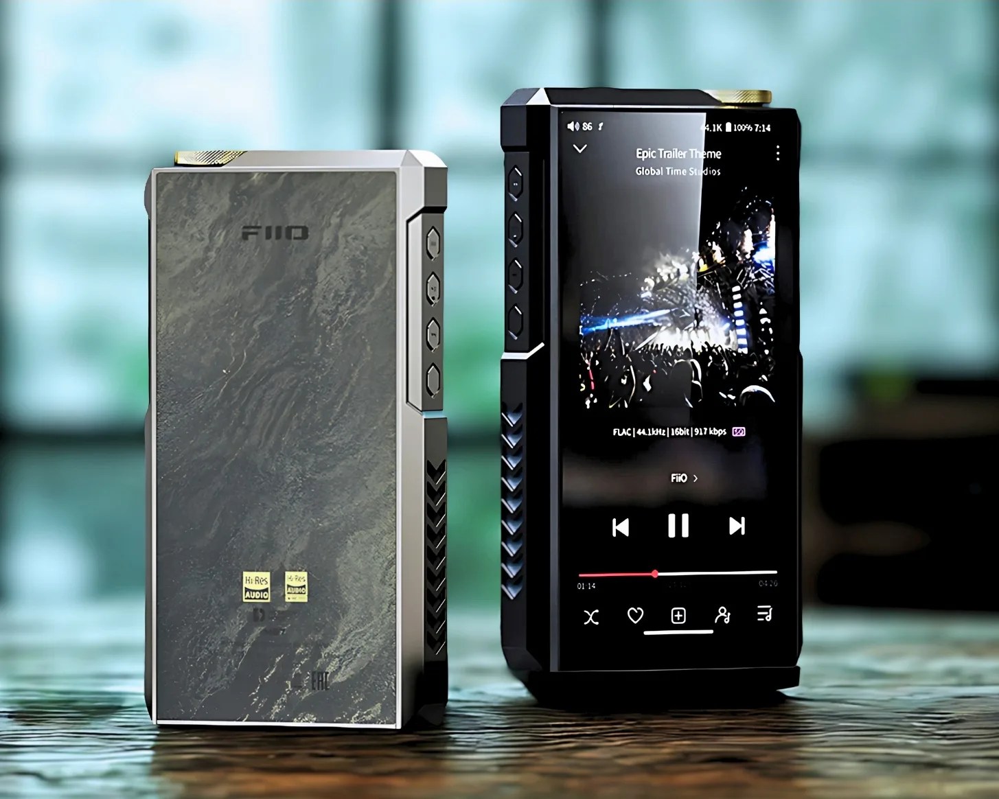 Two portable digital audio players are shown side by side on a reflective surface. The player on the left has a textured, stone-like back panel with the brand "FiiO" and Hi-Res Audio logos. It features four buttons on the side and a gold-colored dial on the top. The player on the right has a glossy black front with a touchscreen displaying a music player interface, showing the track "Epic Trailer Theme" by Global Time Studios. It also has four side buttons and a gold-colored dial on top. Both devices have a modern, angular design with vent-like patterns on the sides.