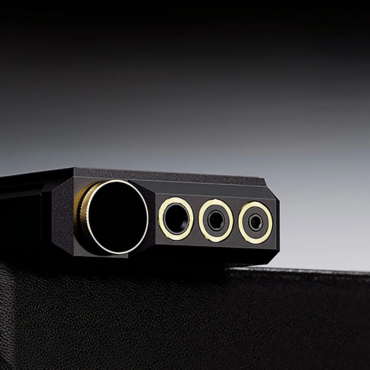 Close-up of a black electronic device featuring a large round knob on the left and three smaller circular ports or connectors with gold-colored rings on the right, set against a gradient gray background. The device rests on a textured black surface.