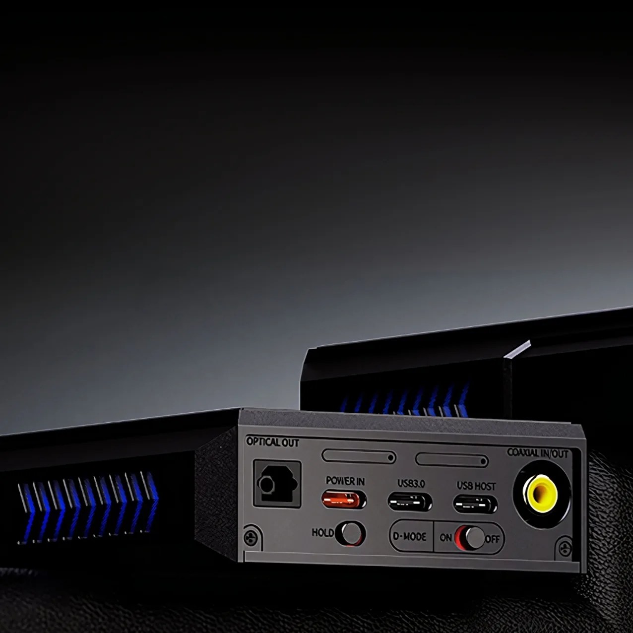 Close-up of the back panel of an electronic device showing various ports and switches. Visible are an optical out port, a power input labeled "POWER IN," two USB-C ports labeled "USB3.0" and "USB HOST," a coaxial in/out port with a yellow connector, and three switches labeled "HOLD," "D-MODE," and "ON/OFF." The device has a black casing with blue ventilation slits.
