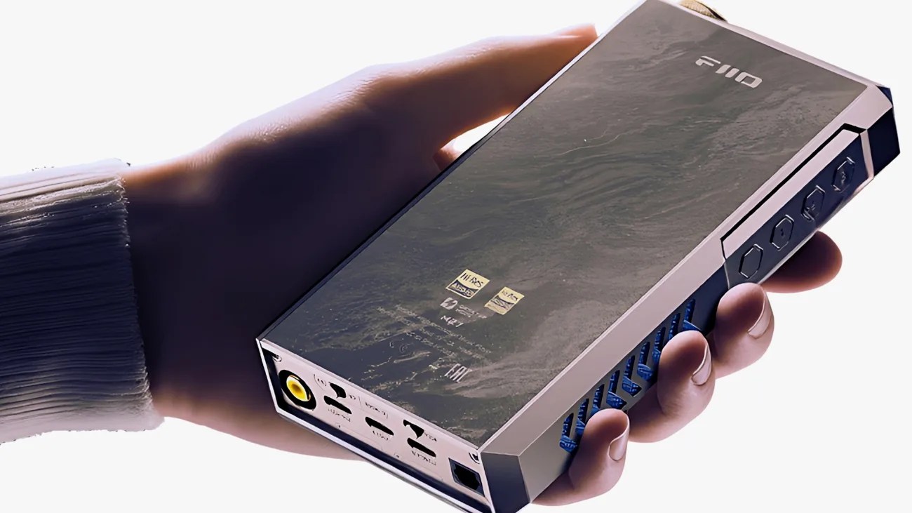 Hand holding a rectangular, metallic audio device with the brand name "FiiO" on the top. The device has multiple ports on one end, including a yellow circular port, and several buttons on the side. The surface has a textured, slightly reflective finish with small logos and text near the center.