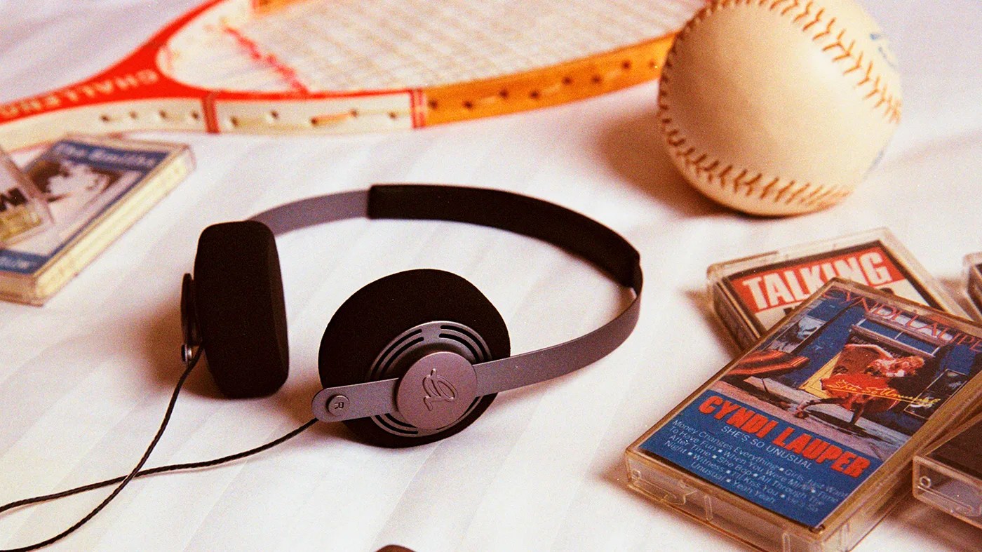 A pair of black and gray over-ear headphones lies on a white surface. Nearby are several cassette tapes, including one with a cover featuring Cyndi Lauper's album "She's So Unusual." In the background, there is a baseball and a tennis racket with an orange frame.