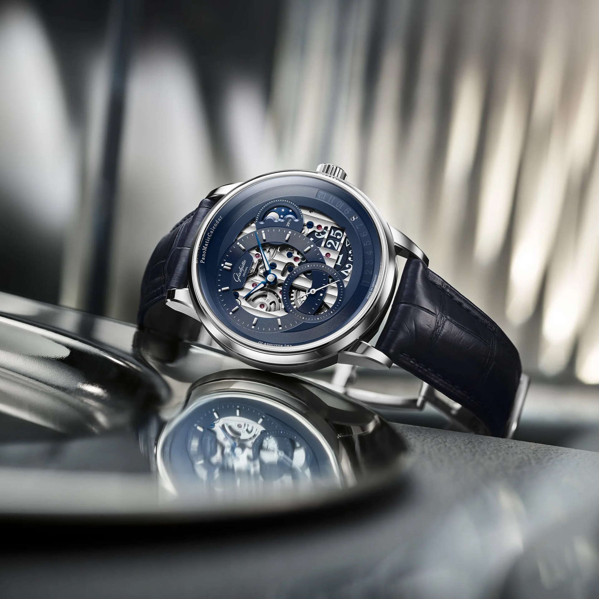 Luxury Glashütte Original wristwatch with a blue skeleton dial showcasing intricate mechanical components. The watch features a moon phase indicator, a date display, and a small seconds subdial. It has a polished silver case and a dark blue leather strap with a textured pattern. The watch is positioned on a reflective surface, creating a mirrored image beneath it.