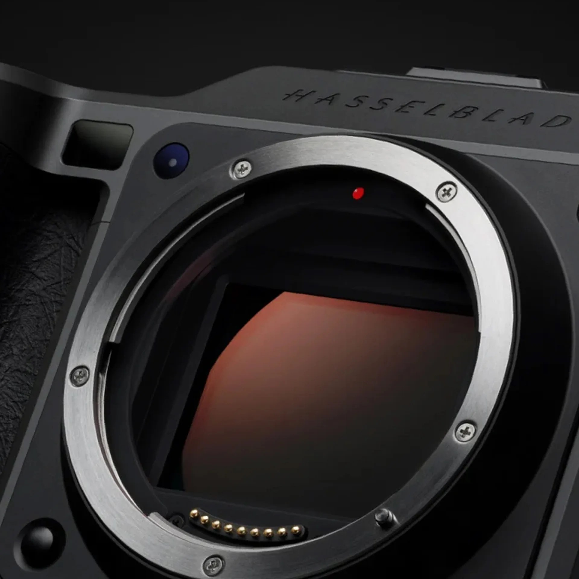 Close-up of a Hasselblad camera body showing the lens mount and sensor inside, with the brand name "HASSELBLAD" engraved on the top. The lens mount is metallic with screws and electronic contacts visible.