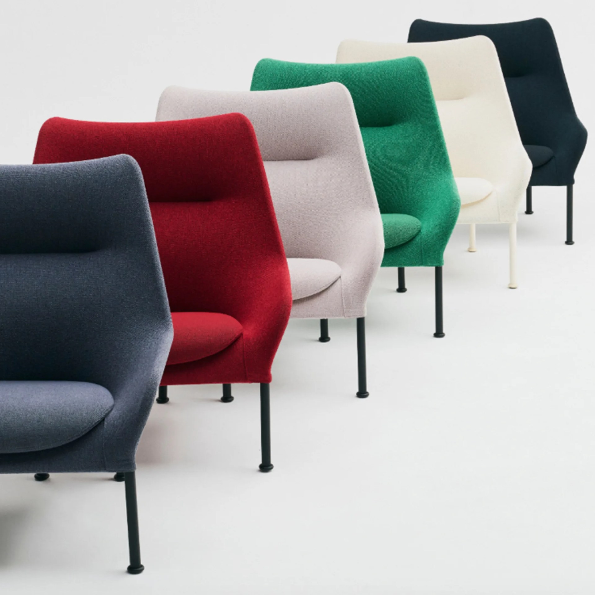 Six modern upholstered hay armchairs in a row, each with a high back and slim black or white legs. The chairs are in different colors from front to back: dark gray, red, light gray, green, off-white, and dark blue. The design is sleek and minimalist, with a slightly curved backrest and a cushioned seat. The background is plain white.
