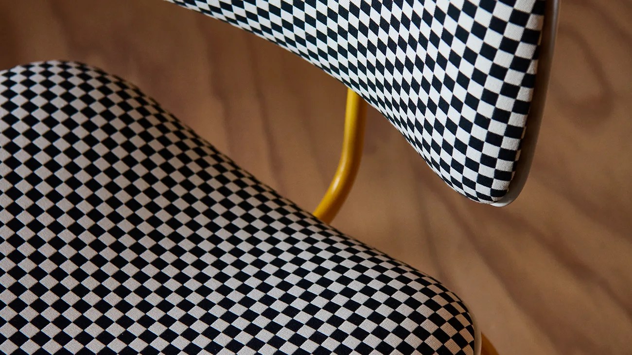 Close-up of a chair with black and white checkered upholstery on the seat and backrest, supported by a yellow metal frame against a brown background.