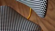Close-up of a chair with black and white checkered upholstery on the seat and backrest, supported by a yellow metal frame against a brown background.