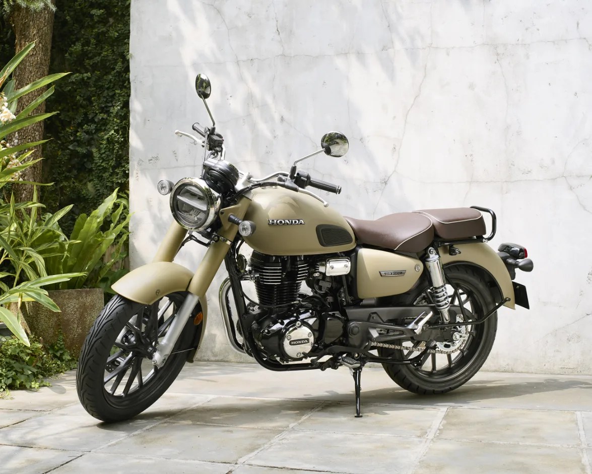 A matte beige Honda motorcycle with a classic design, featuring a round headlight, black engine components, and a brown leather seat. The bike is parked on a tiled surface against a white textured wall with greenery on the left side.