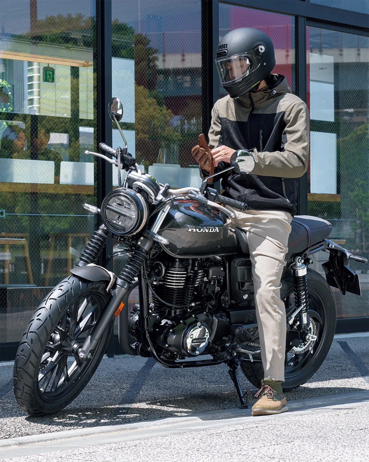 Man wearing a black and beige jacket, beige pants, brown shoes, and a black Shoei motorcycle helmet, sitting on a black Honda motorcycle while putting on brown gloves, parked on a sidewalk in front of a glass building.