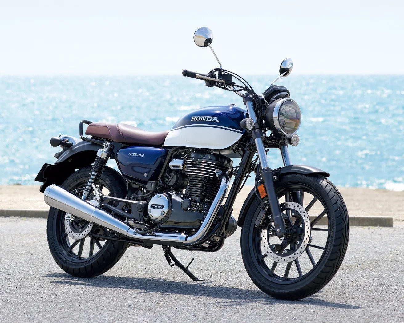 Blue and white Honda GB350 motorcycle with a brown seat, parked on a paved surface near the ocean. The bike features a round headlight, chrome exhaust, and black alloy wheels.