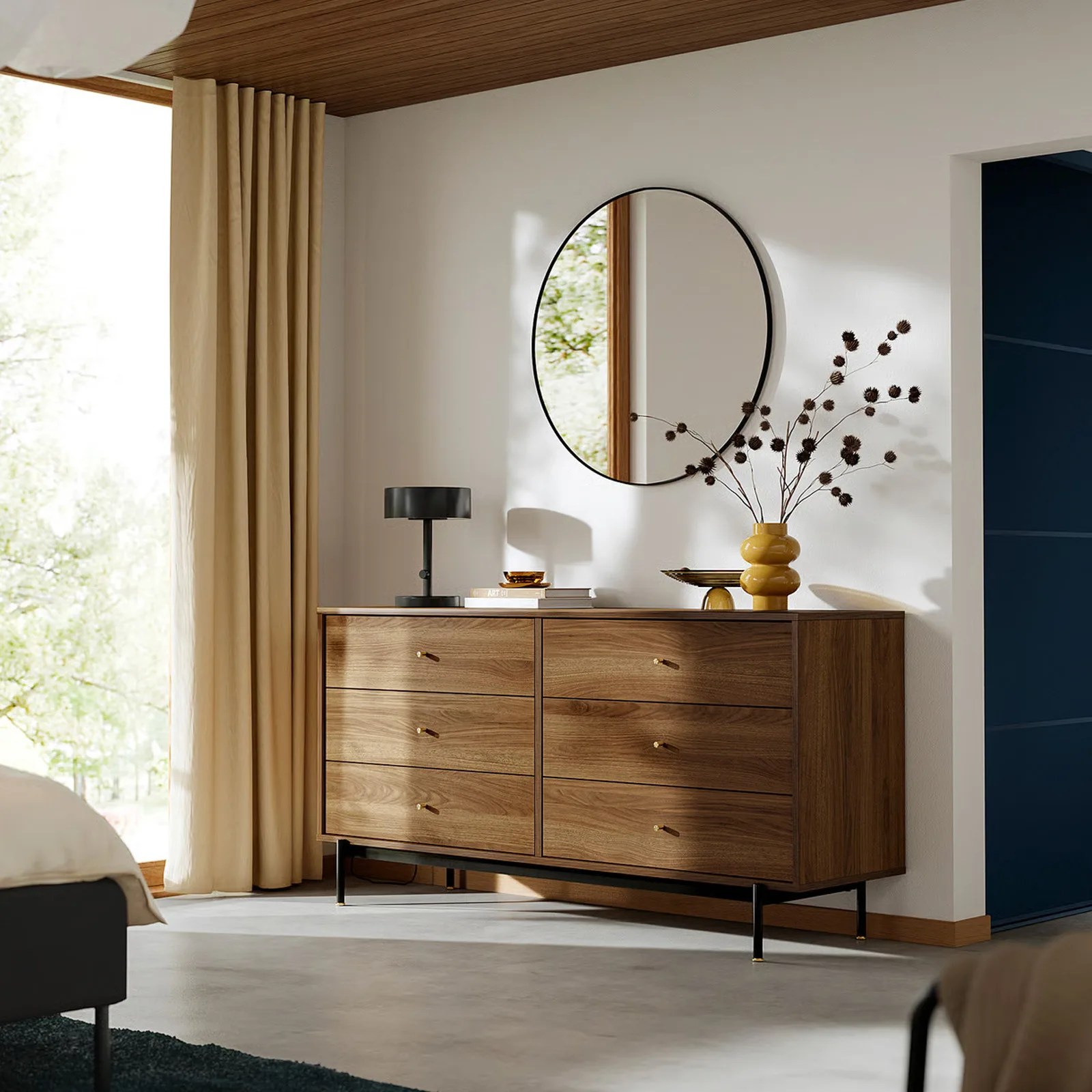 The IKEA RADMANSO six-drawer dresser is shown against a white wall against a floor to ceiling window with the curtains open. A circular mirror is mounted above the dresser alongside a dark table lamp and a vase of flowers