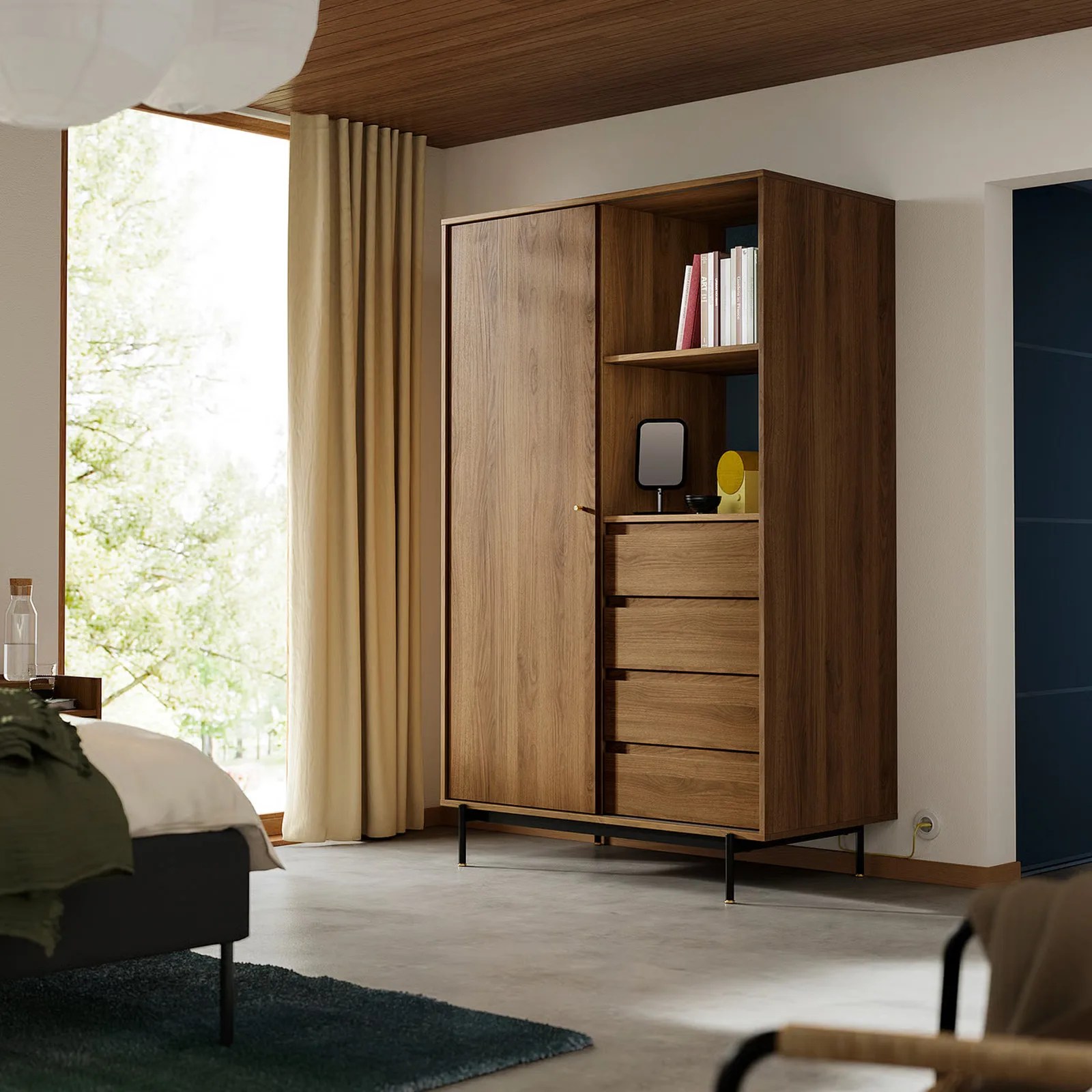 The IKEA RADMANSO wardrobe shown placed in the corner of a bedroom next to a floor to cieling window with the curtains pulled to the side. The dresser is shown holding books along with some other interior design items