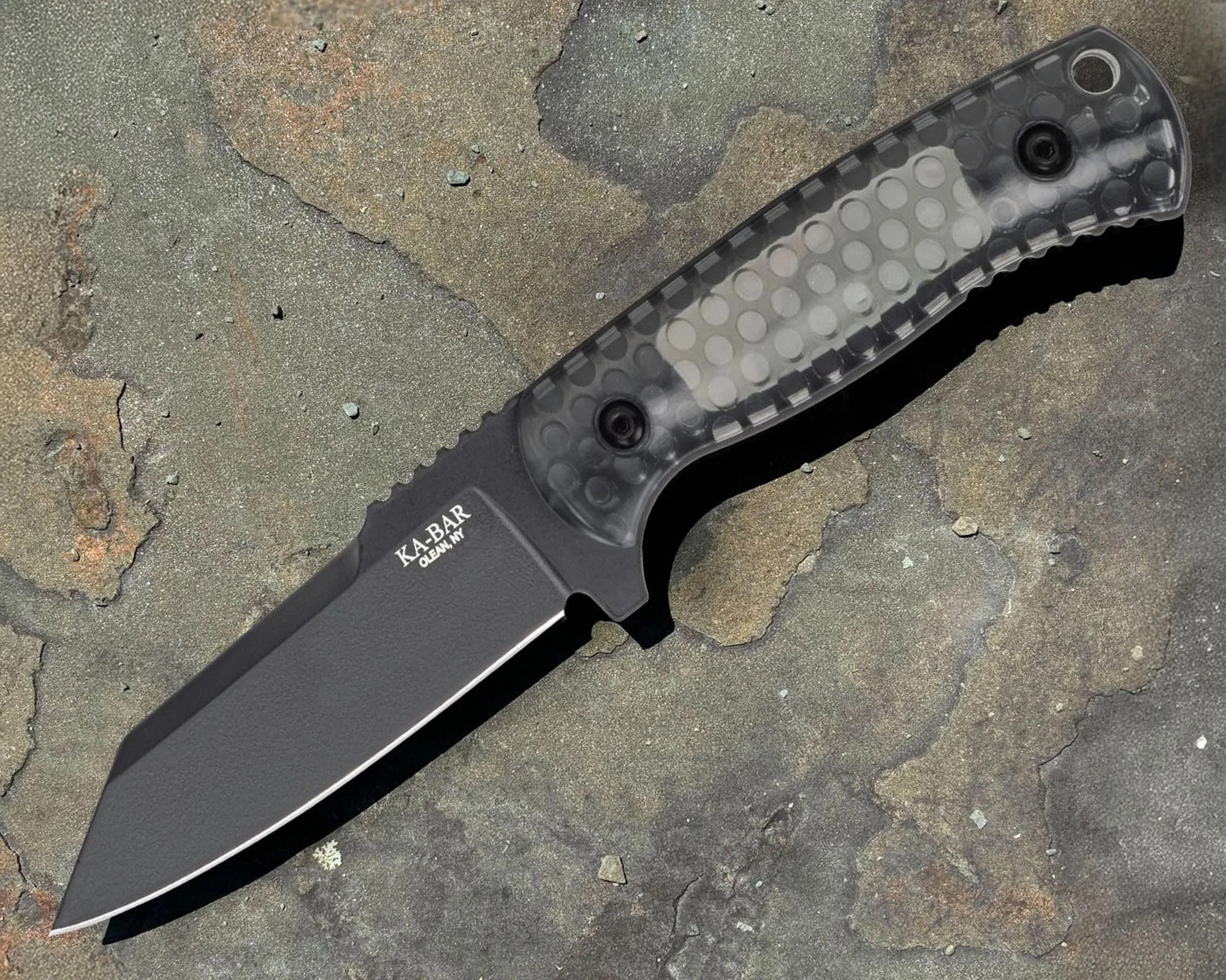 Fixed-blade knife with a black tanto-style blade and a textured, translucent gray handle featuring circular patterns. The blade is marked with "KA-BAR OLEAN, NY." The knife is placed on a rough, multicolored stone surface.