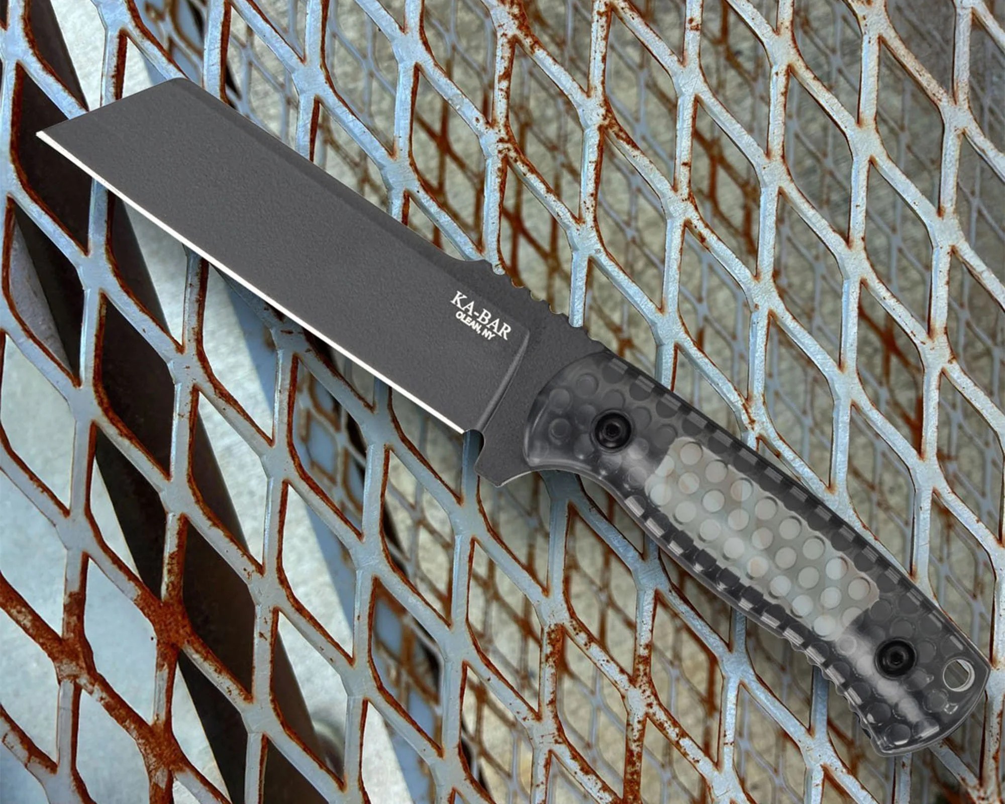 A fixed-blade knife with a black tanto-style blade and a textured, translucent gray handle resting on a rusted metal grid surface. The blade has the text "KA-BAR OLEAN, NY" printed near the handle.