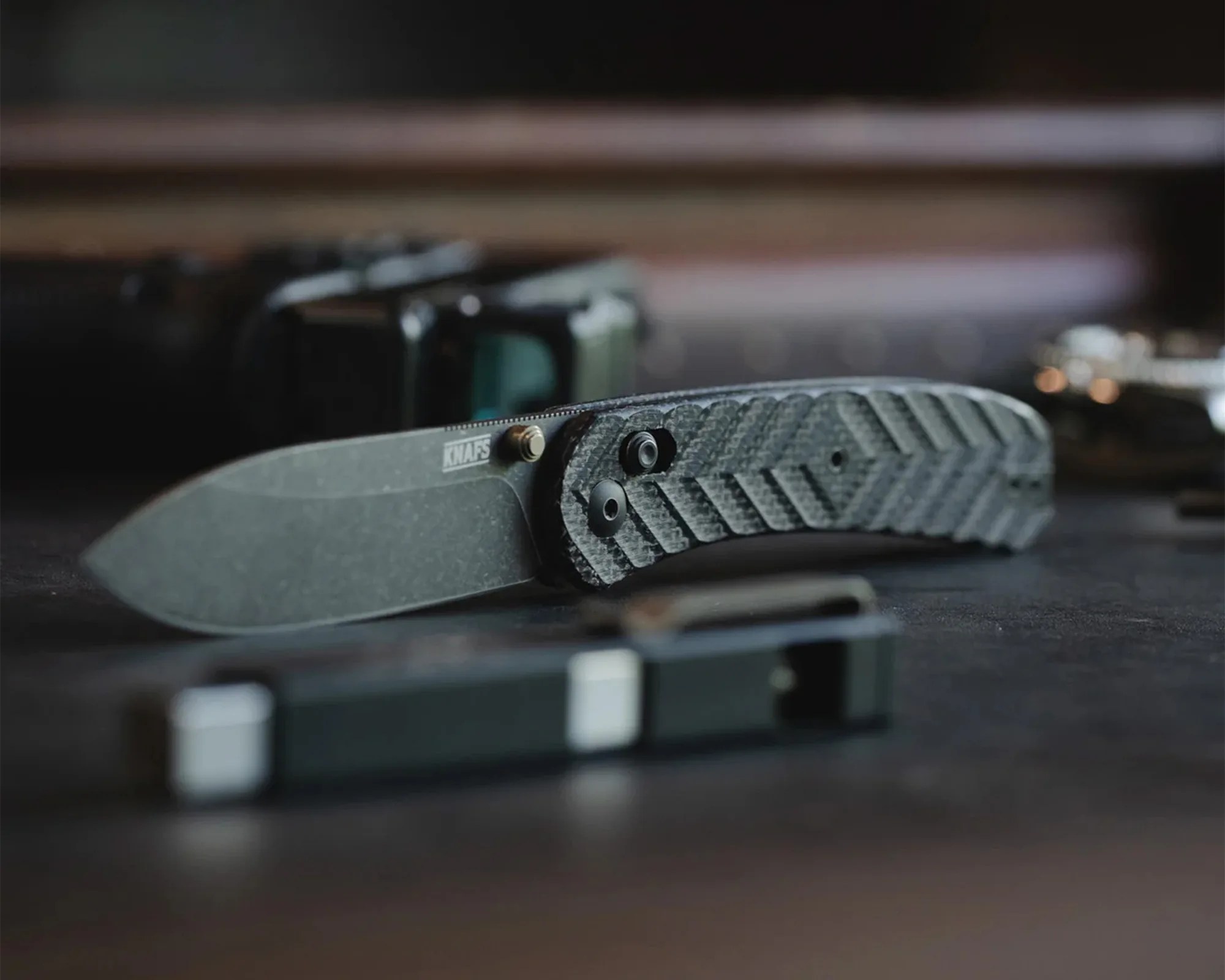A folding knife with a textured, patterned handle and a stonewashed blade. The blade has the brand name "KNAFS" near the handle. The knife is placed on a dark surface with blurred objects in the background.