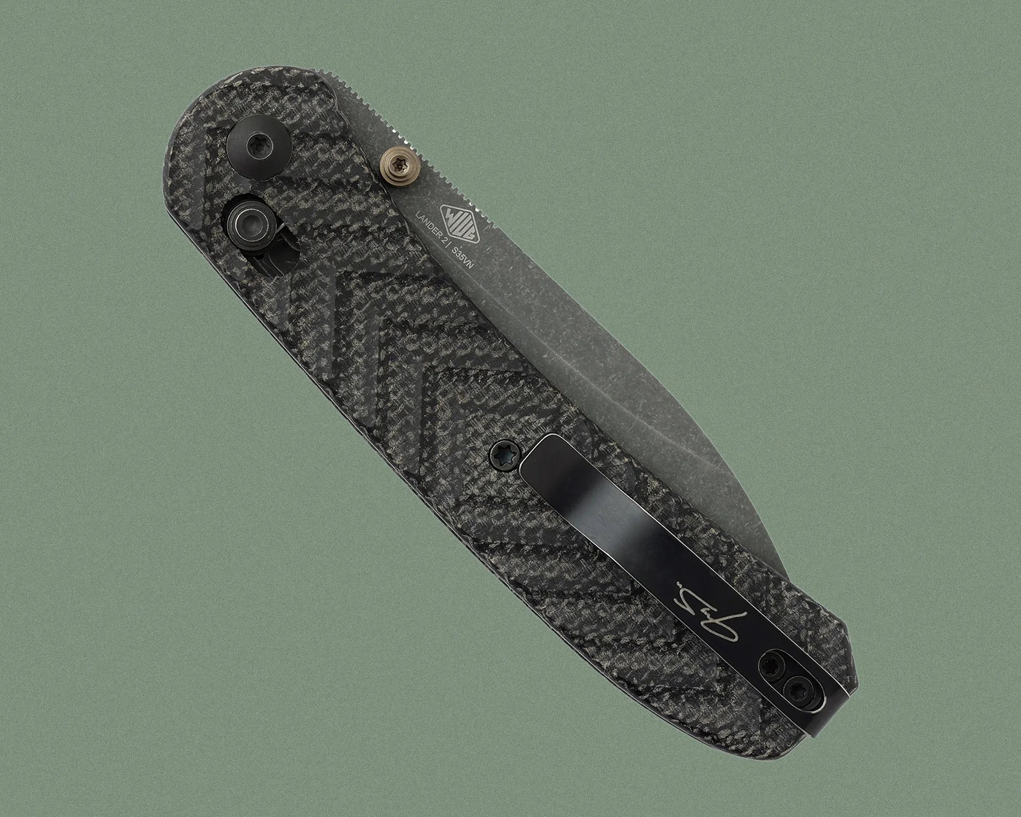 A closed folding knife with a textured black carbon fiber handle featuring a zigzag pattern. The blade is partially visible, made of stonewashed steel, and marked with "LANDER 2 / S35VN" and a logo. The handle has a black pocket clip with a signature or initials on it, secured by two screws. The knife has a thumb stud and a pivot screw near the blade's base.