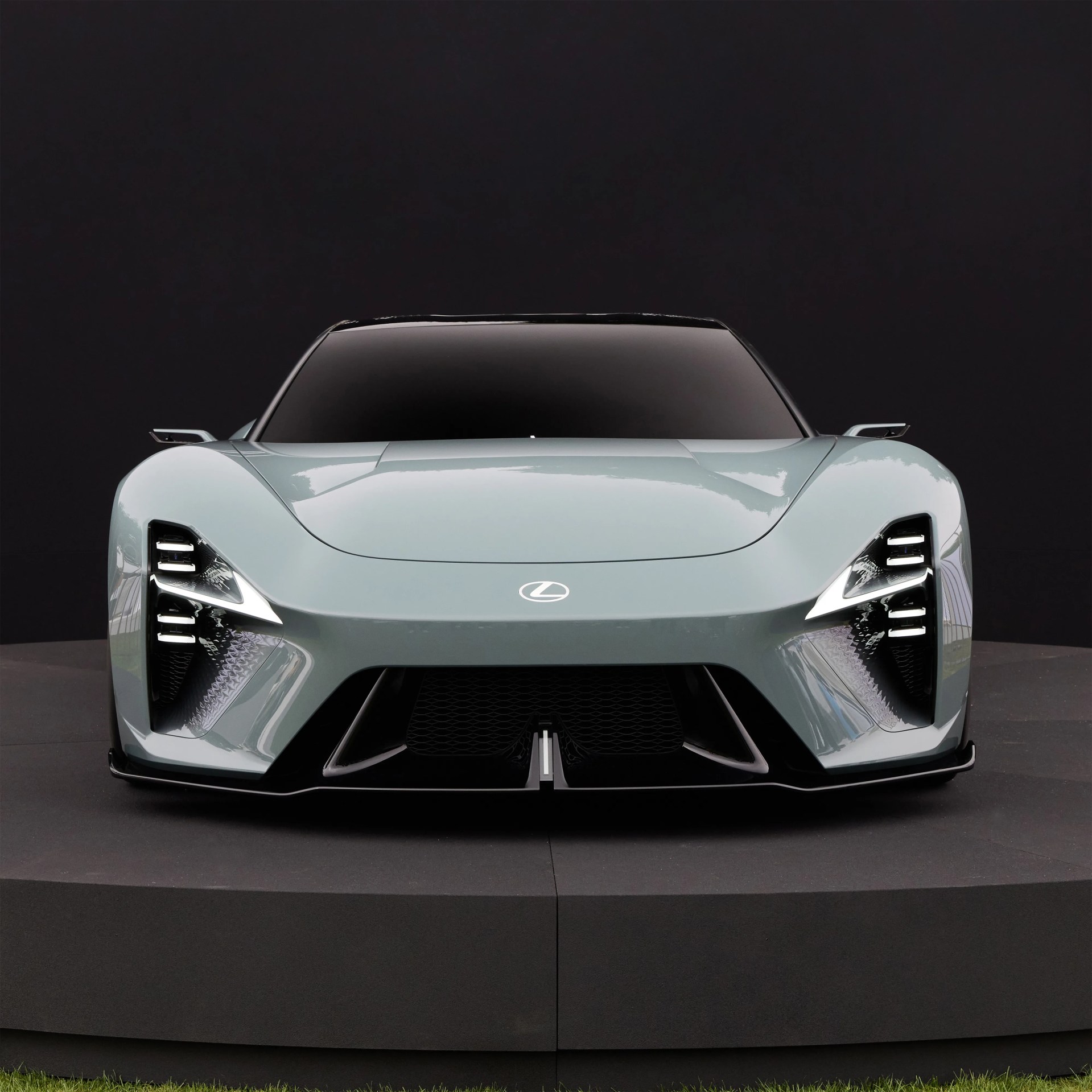 Sleek, futuristic Lexus sports car in a light gray color, shown from the front with a low, wide stance. The car features sharp, angular LED headlights with multiple horizontal light bars on each side, a smooth hood with the Lexus emblem centered, and a large black grille with aerodynamic vents. The windshield is darkly tinted, and the side mirrors are small and streamlined. The car is displayed on a circular black platform against a dark background.