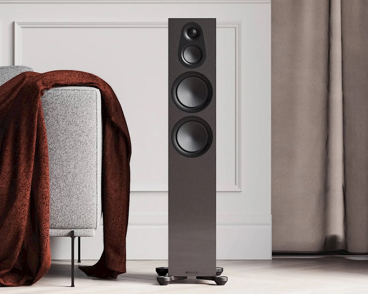 A tall, black floor-standing speaker with three visible drivers (a tweeter, midrange, and woofer) is positioned on a light-colored wooden floor. To the left, there is a light gray upholstered sofa with a dark red blanket draped over its arm. The background features a white wall with decorative molding and beige curtains on the right side.
