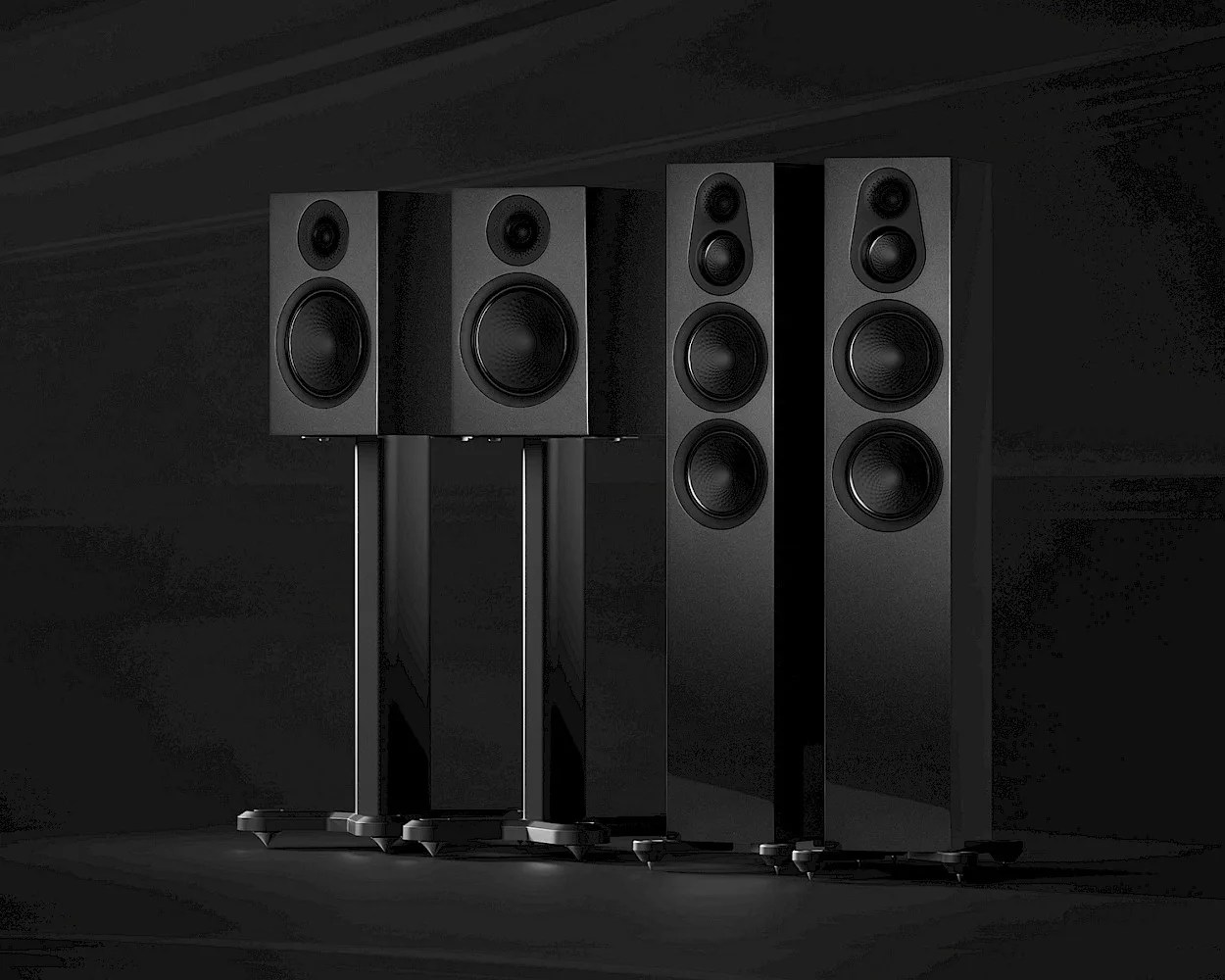 Two pairs of black speakers are shown against a dark background. The pair on the left are bookshelf speakers mounted on tall, slim stands with a tweeter and a larger woofer. The pair on the right are floor-standing speakers with three drivers each: a tweeter, a midrange, and two woofers. All speakers have a sleek, matte black finish.