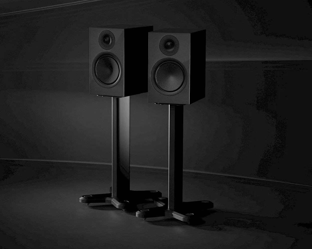 Two black bookshelf speakers mounted on matching black stands, set against a dark background. Each speaker has a circular woofer and a smaller circular tweeter above it. The stands have a sturdy base with four feet for stability.