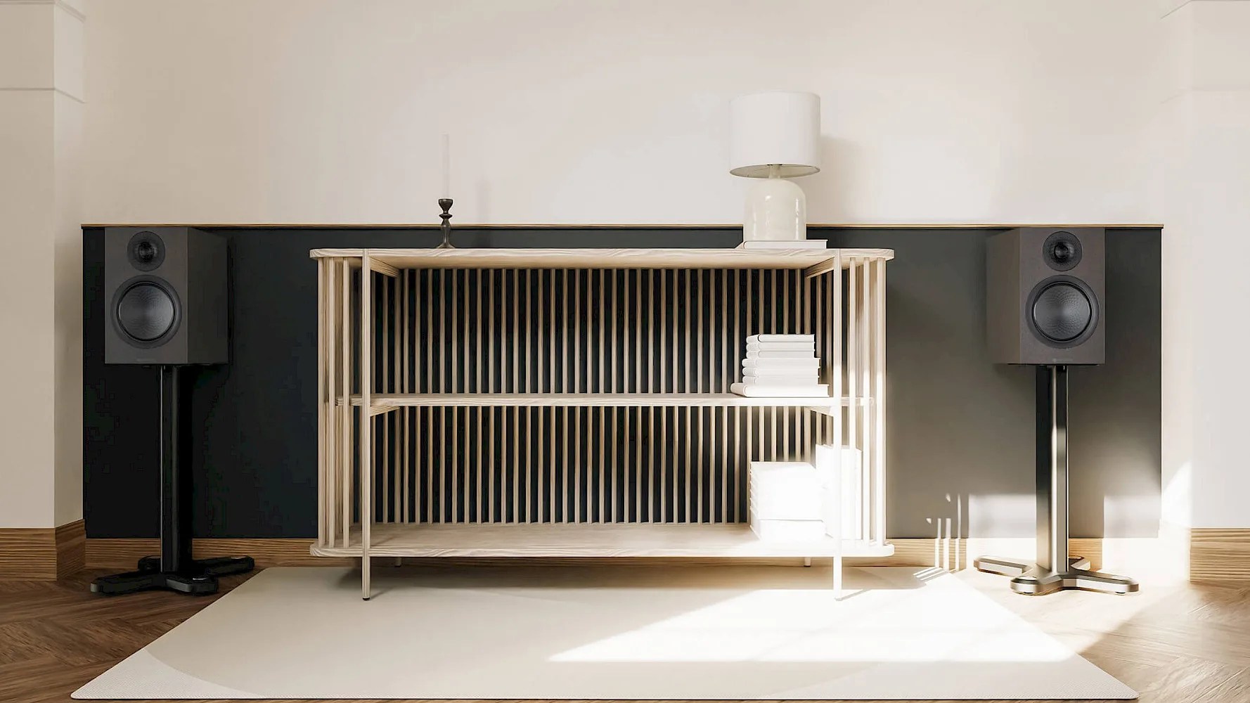 Light wood open shelving unit with vertical slats, holding a stack of white books and a white lamp on top, positioned against a black and white wall. Two black speakers on stands flank the shelving unit, all set on a light-colored rug over a wooden floor.