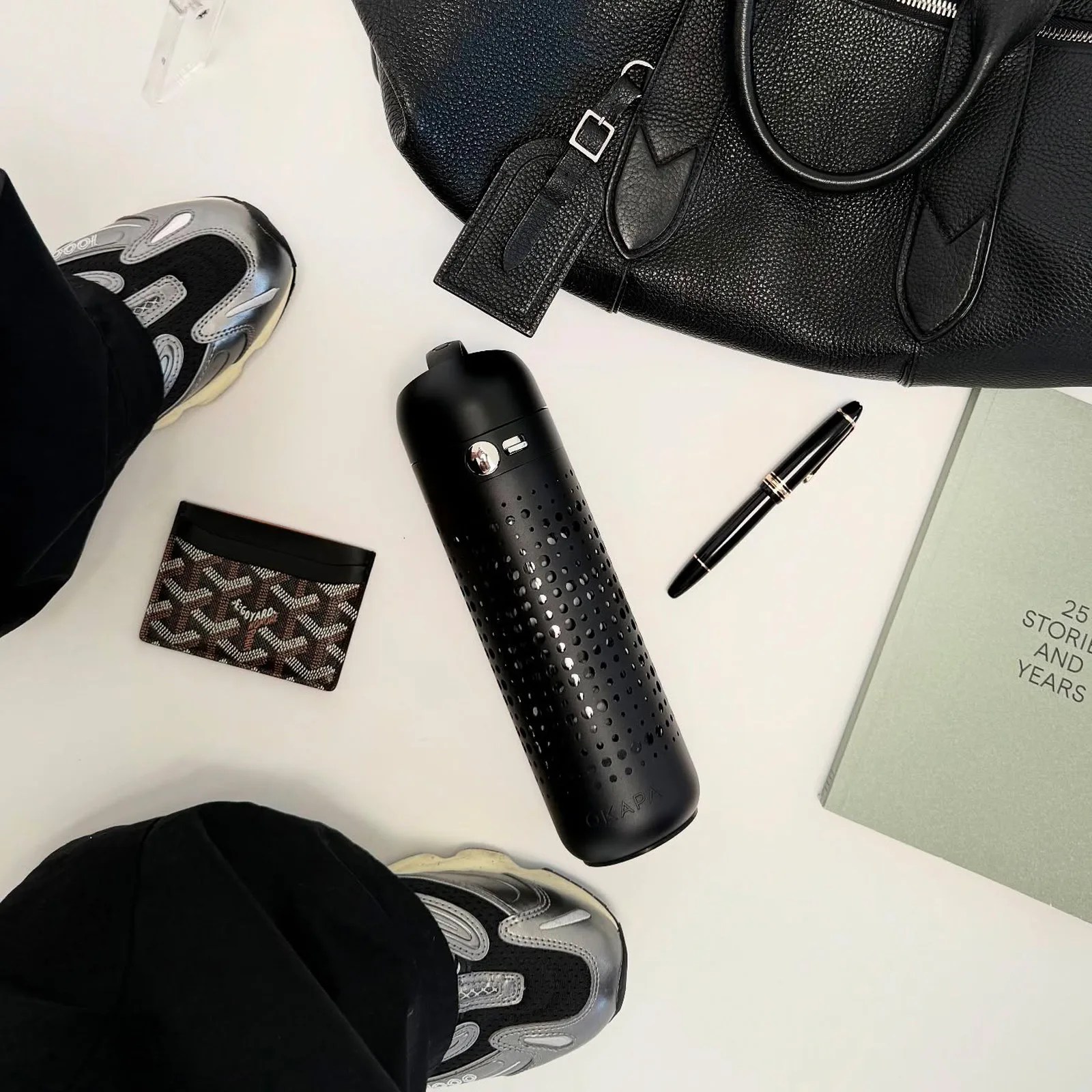 A black perforated cylindrical bottle with a flip-top lid is centered on a white surface. Surrounding it are a black leather handbag with a buckle tag, a black and brown patterned cardholder, a black pen with gold accents, a light green book titled "25 STORIES AND YEARS," and a pair of black and silver sneakers worn by a person dressed in black pants.