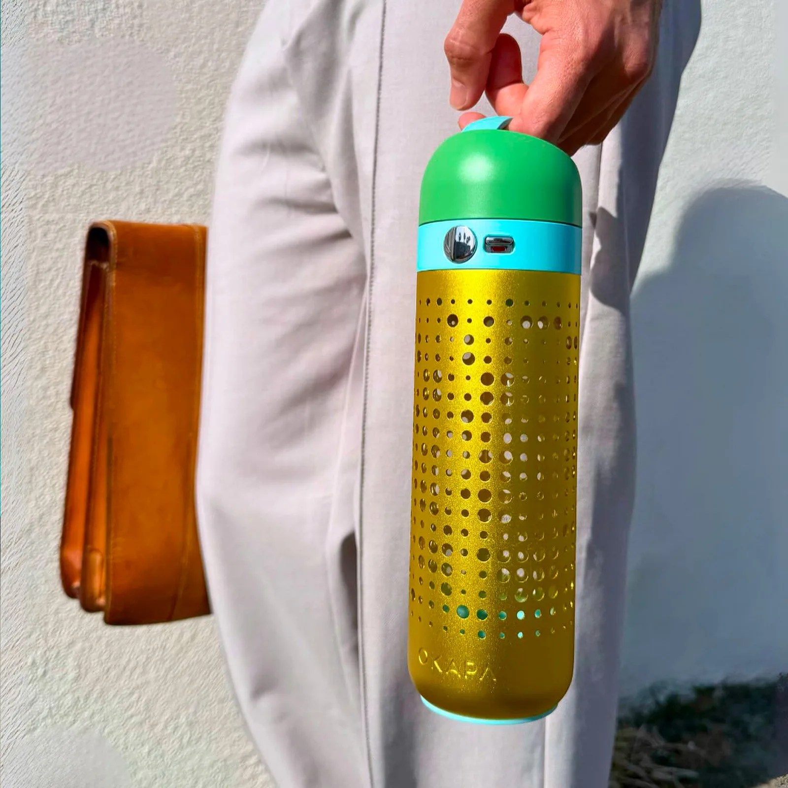 A hand is holding a Okapa with a green top, a blue band near the top featuring a silver button with the brand name "OKAPA" embossed near the bottom. The person is wearing light-colored pants and carrying a brown leather bag.