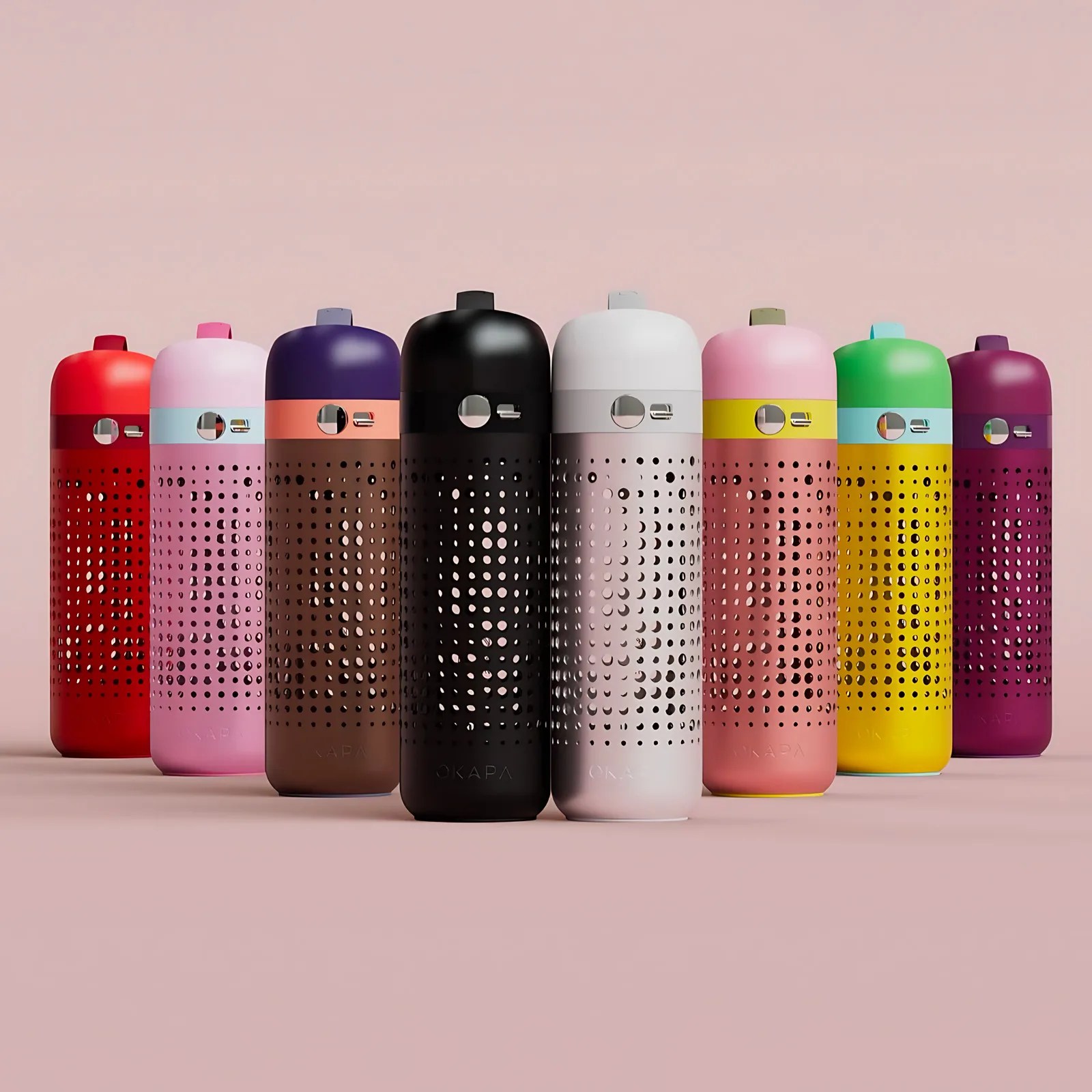 Nine colorful cylindrical Okapa water containers with perforated designs are arranged in a slight arc on a pink surface with a pink background. Each container has a solid-colored top and bottom section with a middle section featuring evenly spaced circular holes. The colors from left to right are red, light pink, dark purple with a peach band, black, white, pink with a yellow band, green with a light blue band, yellow with a light blue band, and dark magenta. Each container has a small circular window and a USB port near the top.
