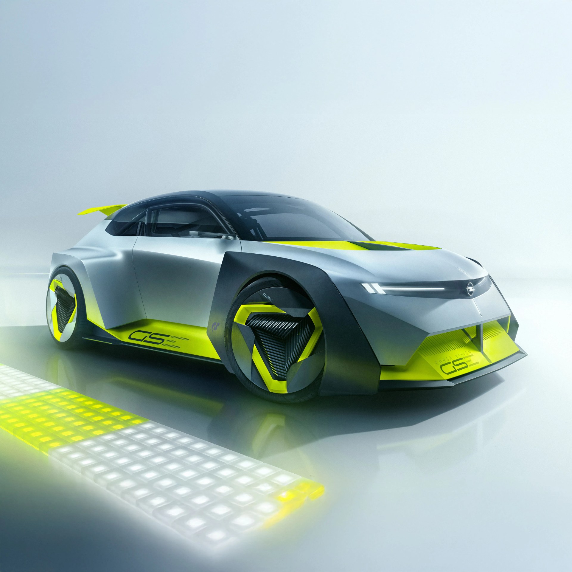 A futuristic Opel concept with a sleek, angular design in silver and black, accented with bright neon yellow highlights. The car features unique triangular wheel covers, a low-profile body, and a prominent rear spoiler. The front has a narrow horizontal light strip and an aggressive, aerodynamic shape. The side of the car displays the text "GSE" in neon yellow. The setting is a minimalistic, reflective surface with soft lighting.