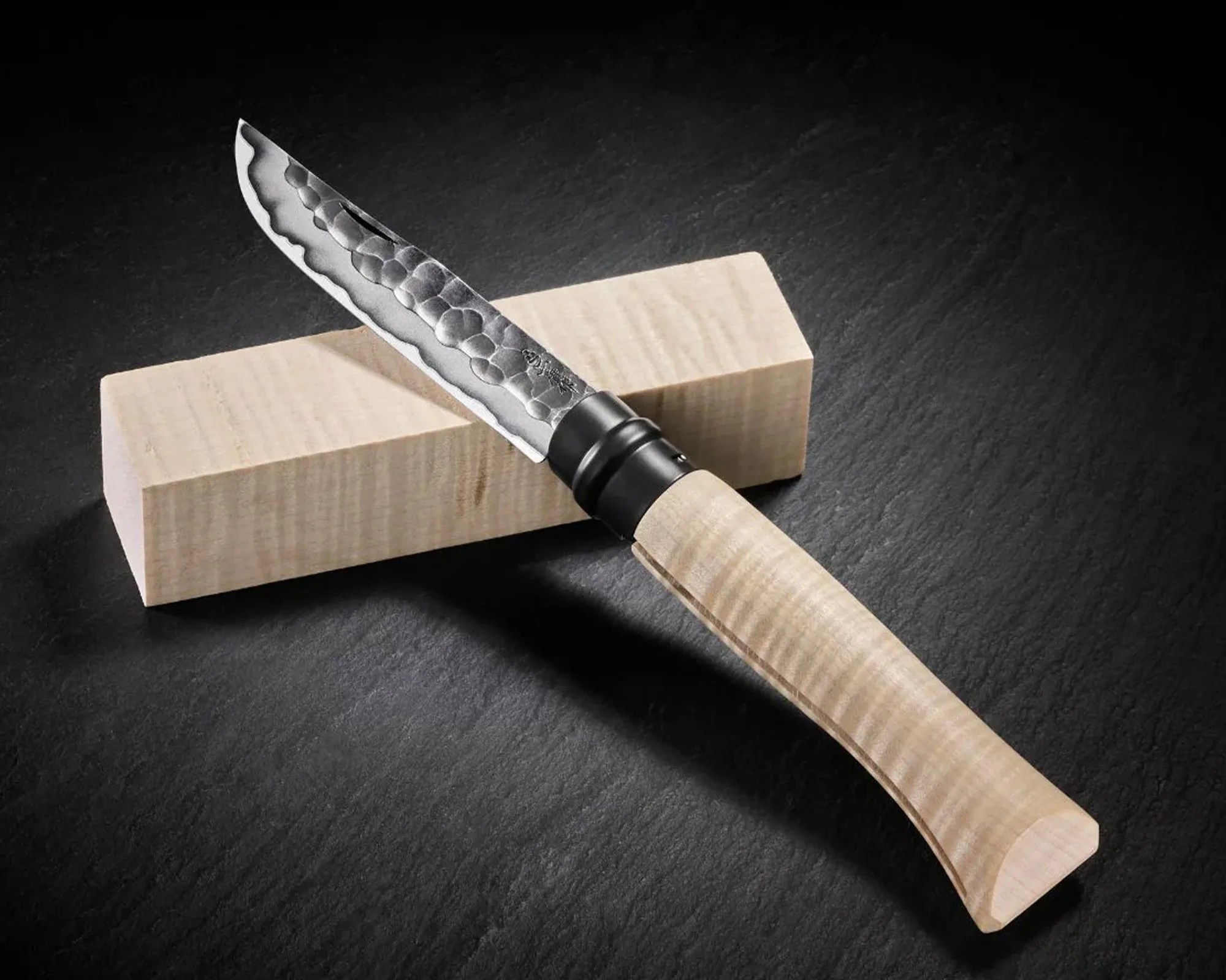 A knife with a hammered, patterned steel blade and a light-colored, textured wooden handle resting on a rectangular block of similar light wood, all placed on a dark textured surface.