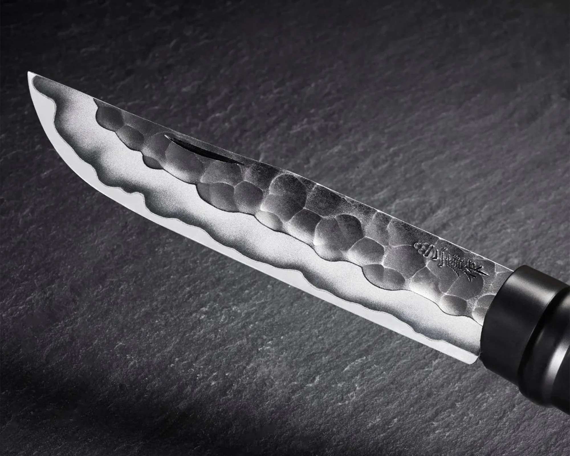 A close-up of a Japanese-style knife blade with a hammered, textured pattern near the spine and a sharp, polished edge. The blade features engraved Japanese characters and has a black handle visible on the right side. The background is a dark, textured surface.