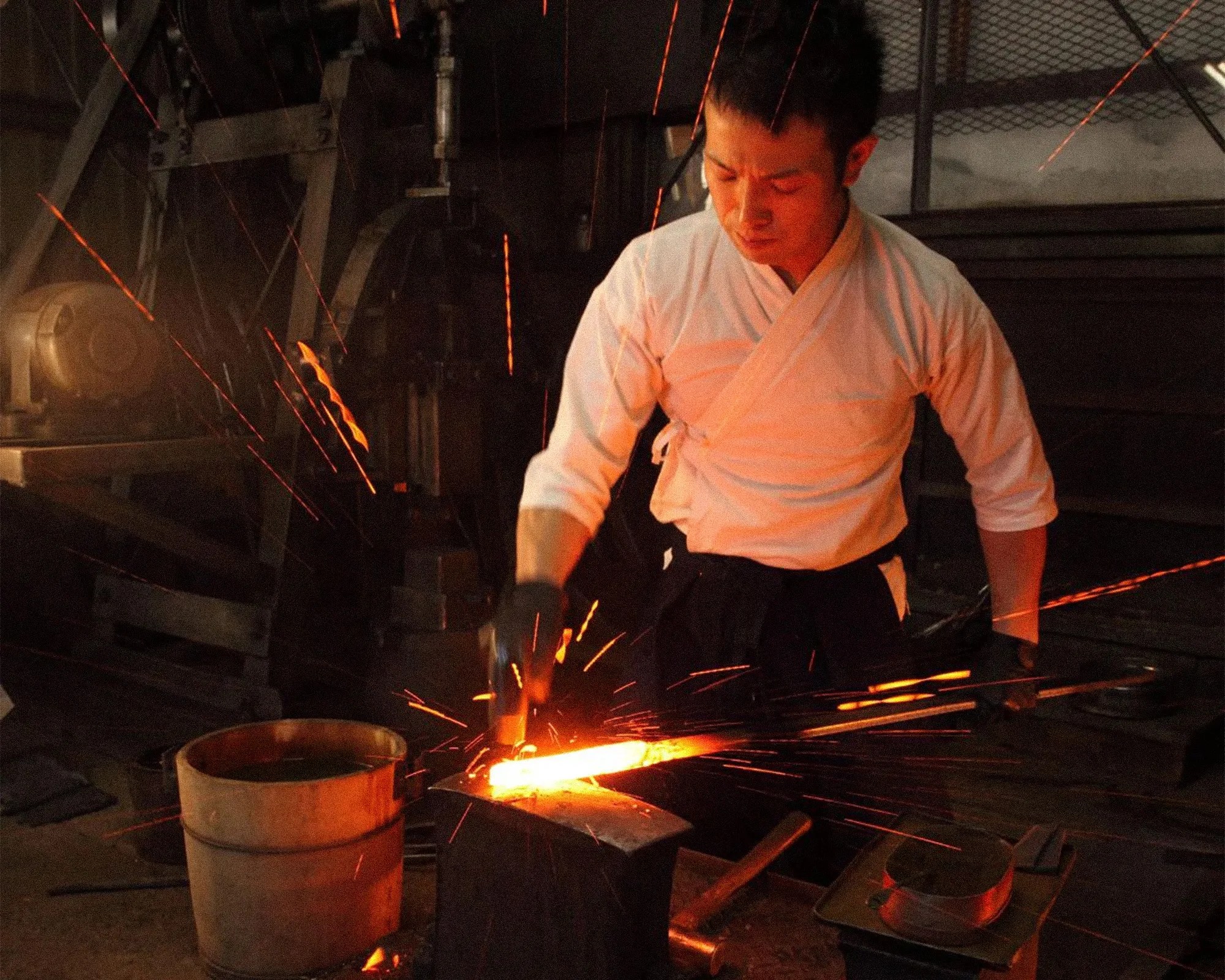 A blacksmith wearing a white traditional shirt and black gloves is hammering a glowing hot piece of metal on an anvil, causing sparks to fly around. The workshop is dimly lit with industrial equipment and tools visible in the background.