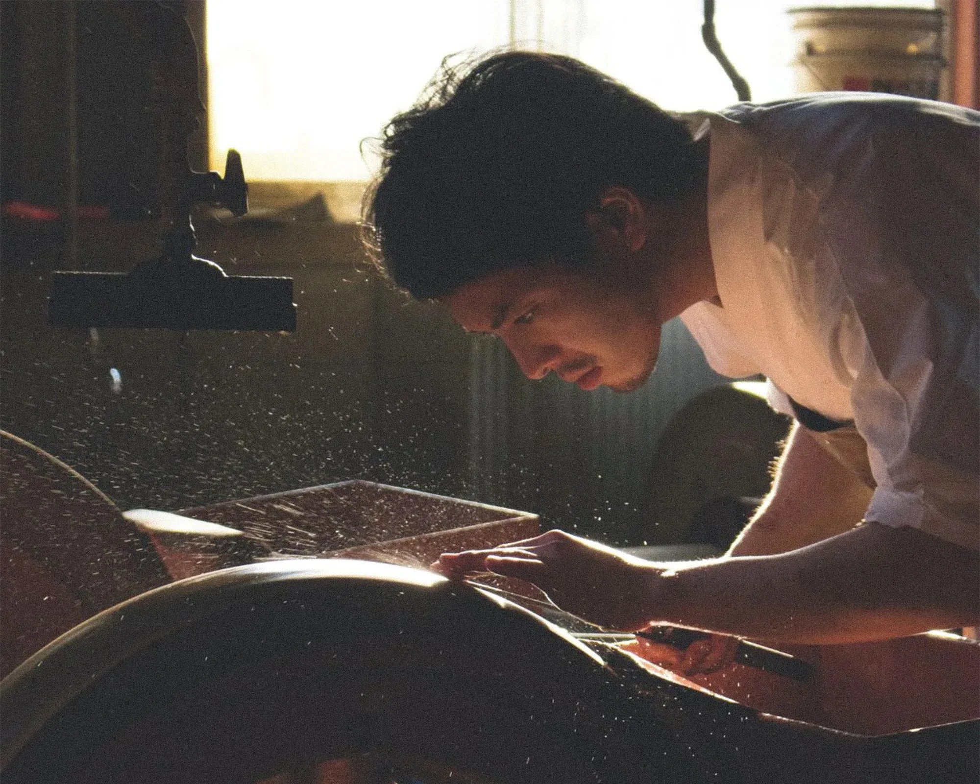 A man is closely inspecting or working on a wooden object, possibly shaping or sanding it. He is leaning forward with intense focus, and wood dust particles are visible in the air, illuminated by soft, warm light coming from a window behind him. The setting appears to be a workshop with woodworking tools and equipment.