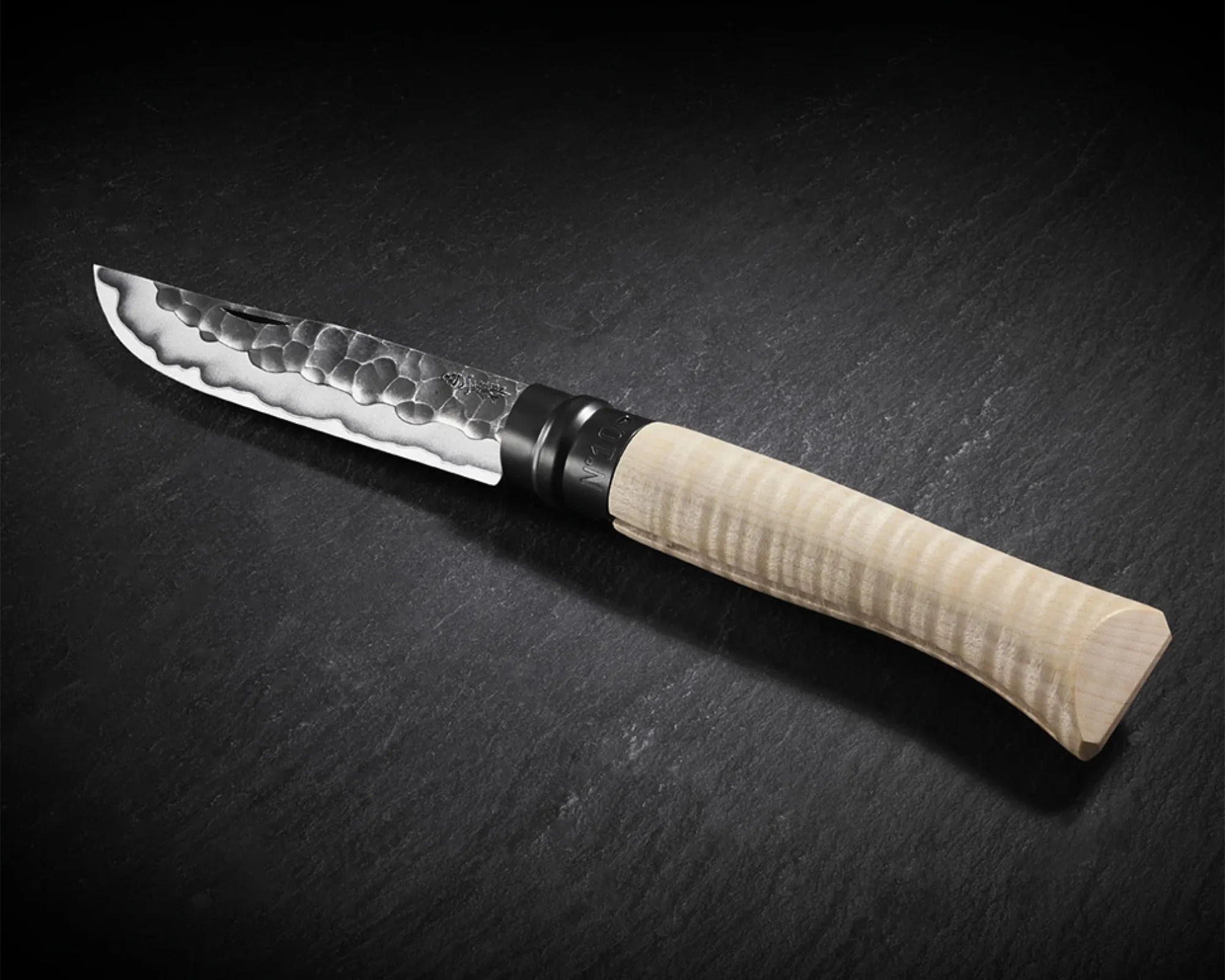 A knife with a hammered, patterned steel blade and a light-colored, textured wooden handle resting on a dark surface. The blade has a wavy edge design near the cutting edge and a small notch near the handle.