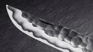 Close-up of a knife blade showing a distinctive hamon pattern along the edge and a hammered texture on the upper part of the blade. The blade has a sharp, pointed tip and a polished cutting edge. The background is dark and textured.