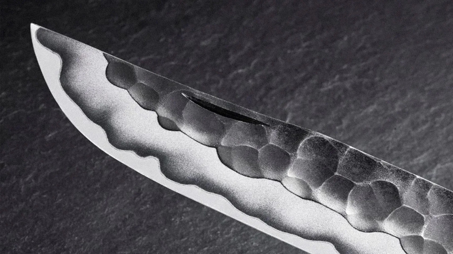 Close-up of a knife blade showing a distinctive hamon pattern along the edge and a hammered texture on the upper part of the blade. The blade has a sharp, pointed tip and a polished cutting edge. The background is dark and textured.