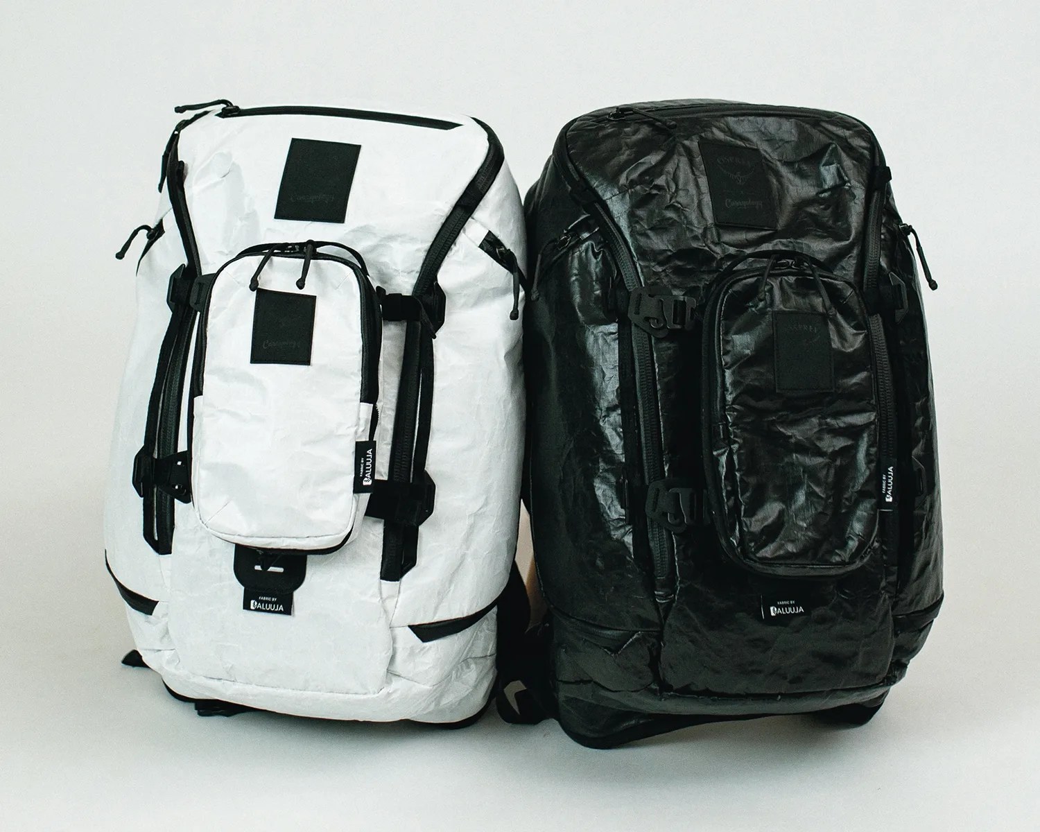 Two large backpacks, one white and one black, made from a crinkled, textured material. Each backpack features multiple zippered compartments, including a prominent front pocket with black zipper pulls and black rectangular patches. Both have black straps and buckles on the sides and a small black label near the bottom that reads "FABRIC BY ALUUJA." The backpacks are standing upright against a plain white background.
