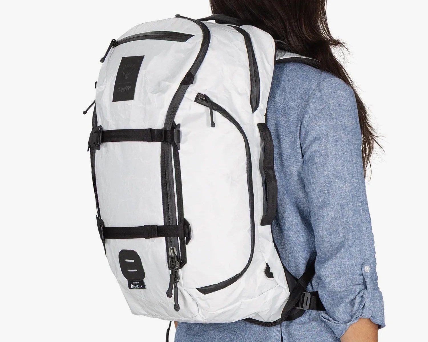 White backpack with black zippers, straps, and accents, worn by a person in a blue long-sleeve shirt. The backpack features multiple compartments, adjustable side straps, and a black patch near the top.