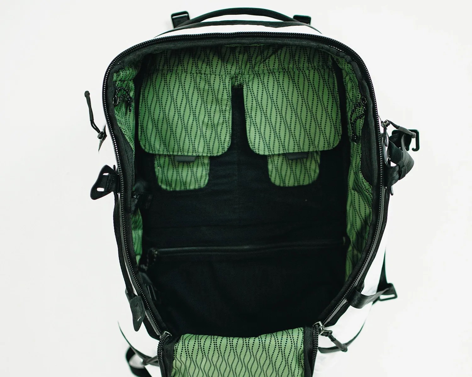 Open black backpack with a green interior lining featuring a wavy dotted pattern. Inside, there are two flap pockets at the top and a zippered pocket at the bottom. The backpack has black zippers and straps.