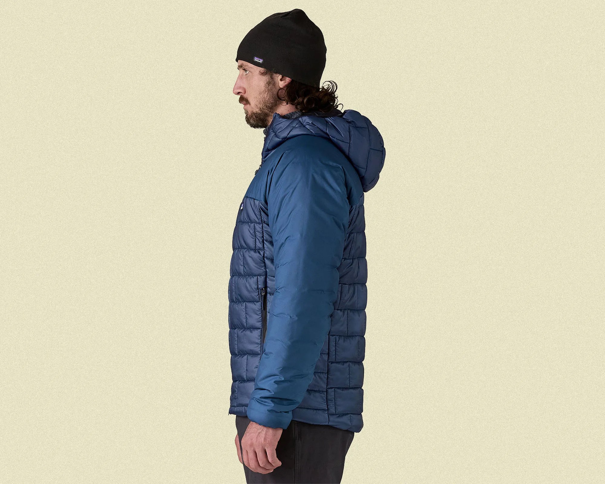 Man wearing a blue quilted jacket with a hood and a black beanie, shown in profile against a plain light beige background.