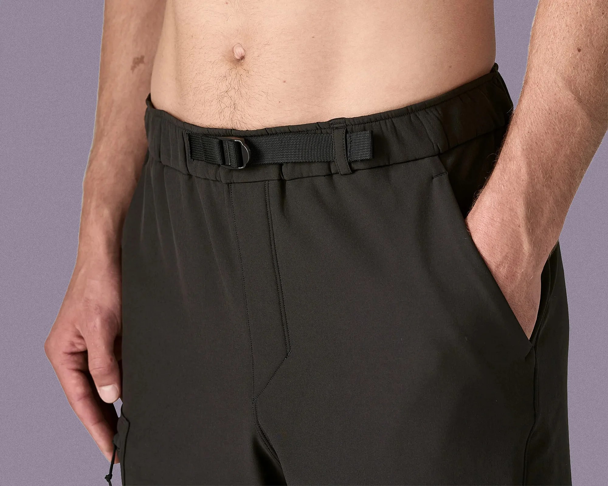 Close-up of a person wearing black pants with an integrated black adjustable belt and elastic waistband. The person's left hand is in the left pocket, and the right hand is relaxed by their side. The background is a solid muted purple color.