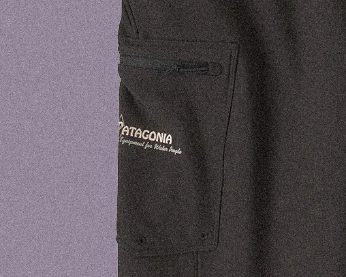 Close-up of a black Patagonia garment pocket with a horizontal zipper and the Patagonia logo printed in white, along with the text "Equipment for Whole People." The pocket has two small black rivets at the bottom corners.