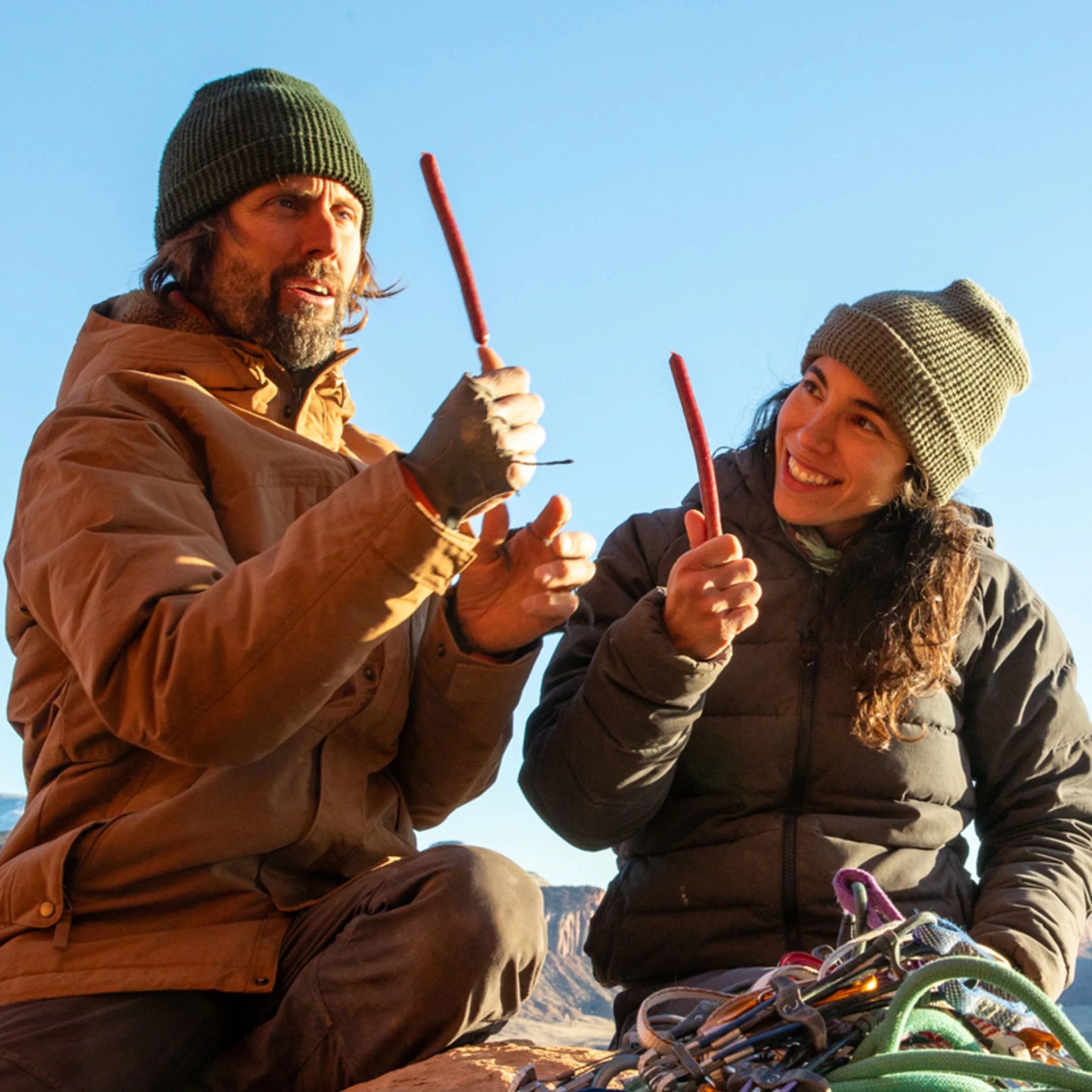 Two people dressed in warm outdoor clothing and knit hats are sitting on a rock, each holding a stick of jerky. They are smiling and appear to be enjoying a snack together, with climbing gear including ropes and carabiners visible in front of them. The background shows a clear blue sky and a rocky landscape.