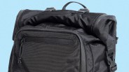 Black roll-top backpack with a front zippered pocket, a top handle, and adjustable straps with plastic buckles, set against a light blue background.
