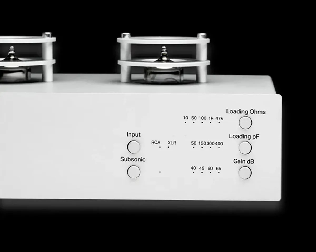 Close-up of a white audio device front panel featuring two circular knobs on the left labeled "Input" with options RCA and XLR, and "Subsonic." On the right side, there are three circular buttons labeled "Loading Ohms," "Loading pF," and "Gain dB," each with corresponding numeric indicators above them. The top of the device shows two metal components with protective white frames.